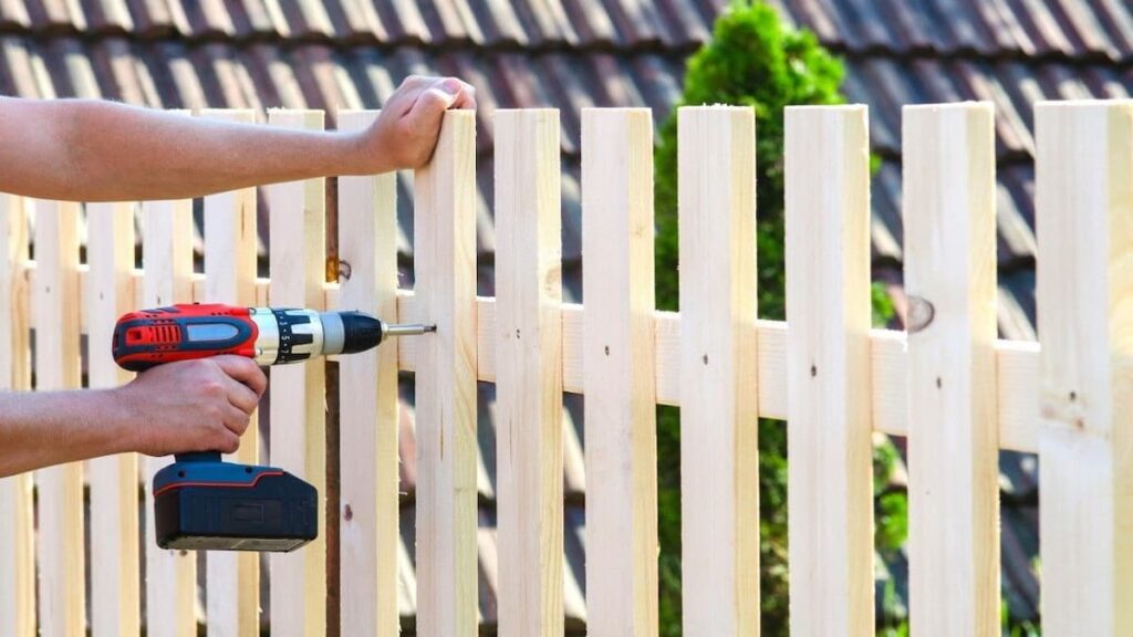 A man drills into wood to install a fence, using countersunk coach screws for secure attachment.