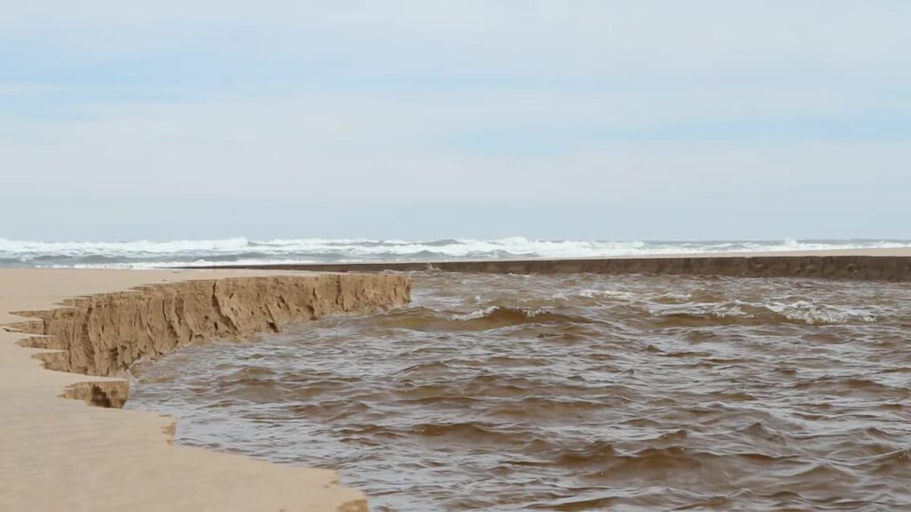 A sand dune erodes as ocean waves crash against it, highlighting the riverbank collapse at Iford Playing Fields.riverbank collapse iford playing fields