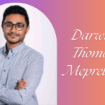 Portrait of Darwin Thomas Meprethu, a middle-aged man with short black hair, wearing glasses and a blue shirt, smiling.