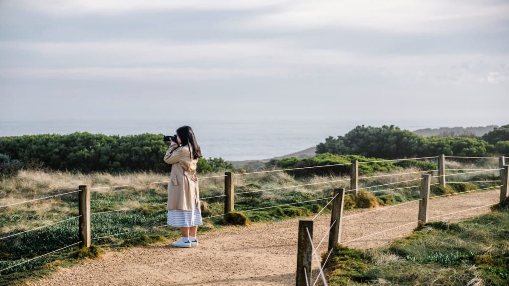 A woman in a trench coat stands on a path by the ocean, with waves gently crashing in the background. beliktal