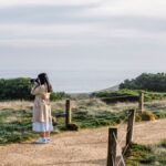 A woman in a trench coat stands on a path by the ocean, with waves gently crashing in the background. beliktal