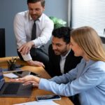Three professionals in business attire collaborate on a laptop during a software meetshaxs update meeting.
