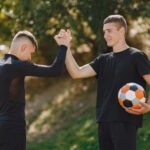 Two young men giving each other a high five while holding a soccer ball, celebrating a successful play together bảce